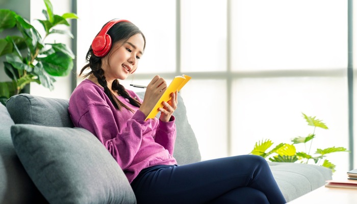 Candid of asian woman listening podcast to learning online by red headphone. Asian pretty girl enjoy and relax in weekend lifestyle on sofa at home.