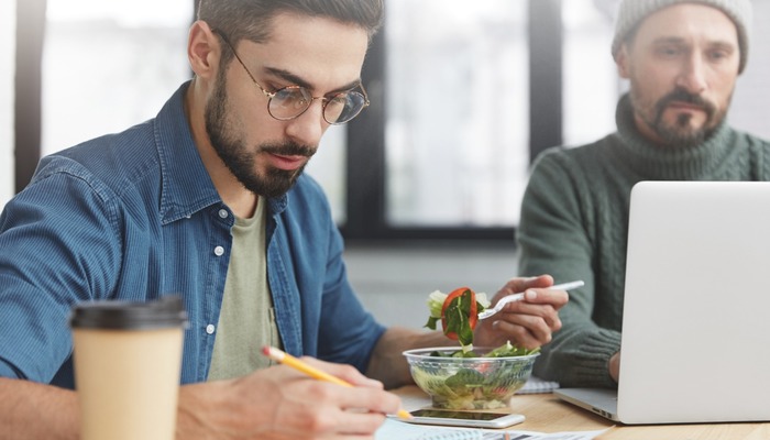 Two corporative workers work together at workplace: mature man keyboards on laptop and his companion writes attentively notes, eats fresh salad and drink coffee, have no time for going in cafe
